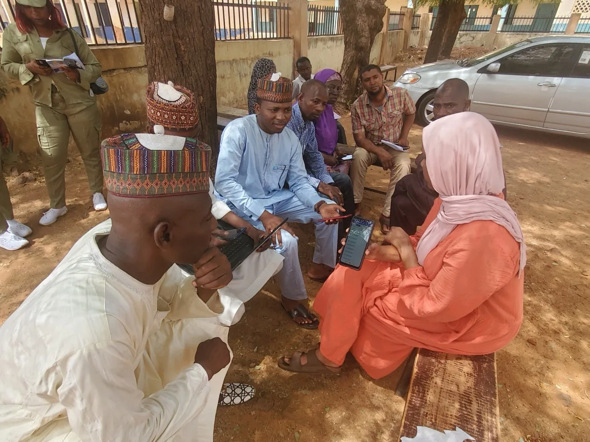 A field campaign manager enrolls a health worker on a mobile phone as community members look on