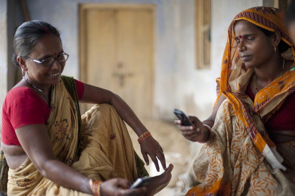 Two women in saris sitting together outdoors, each looking at a mobile phone.