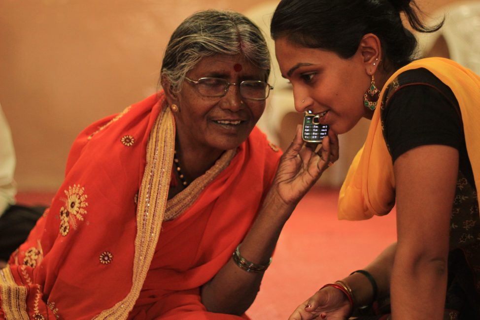 A young woman holding a basic mobile phone to an older woman's ear as the two sit together.