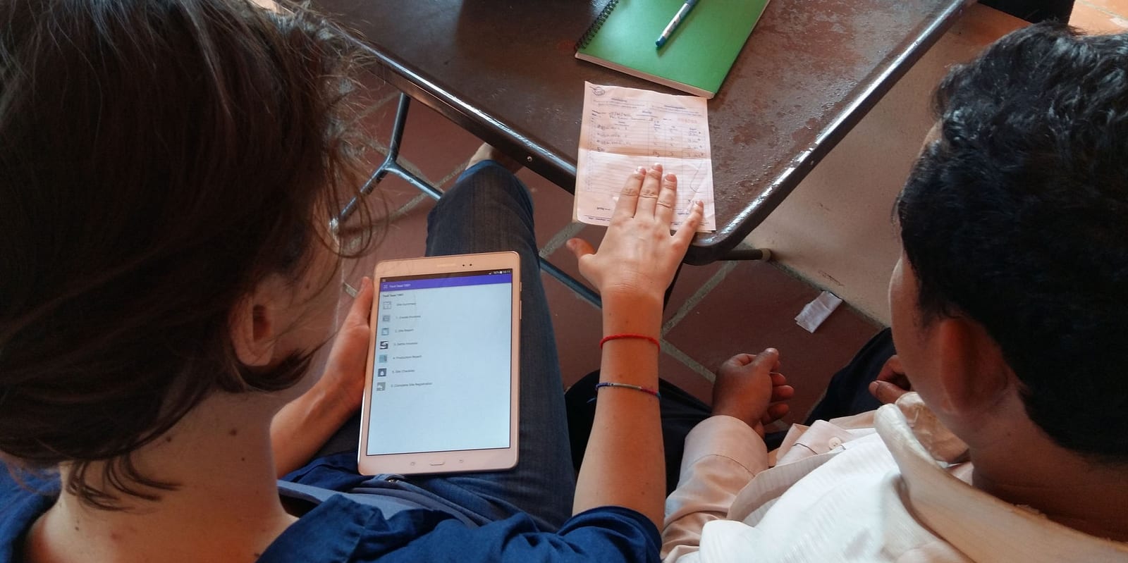 Seen from above, a woman holds a tablet showing a CommCare app menu while sitting beside a colleague and a handwritten paper form on a table.