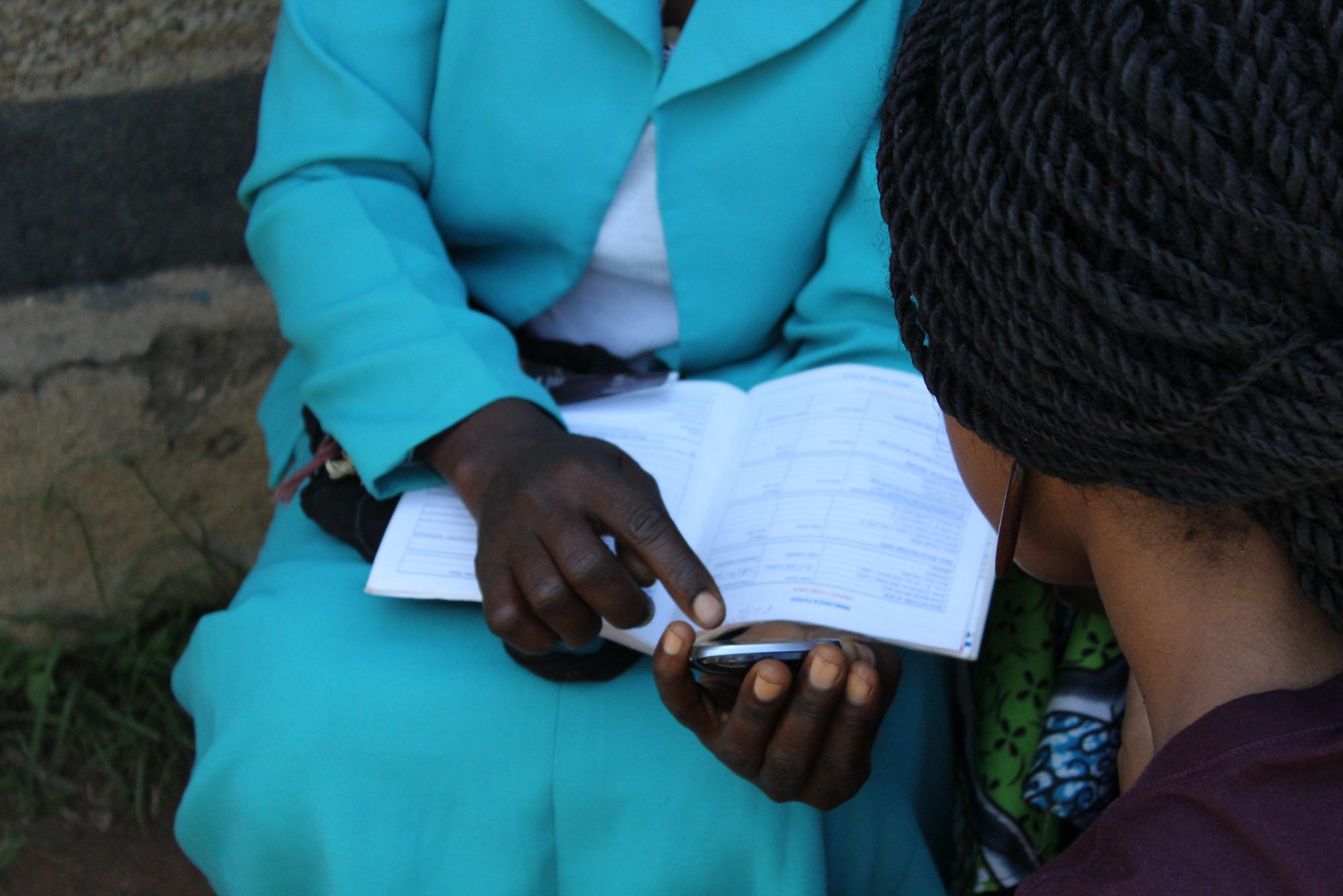 A health worker cross-referencing a paper patient register with a basic mobile phone