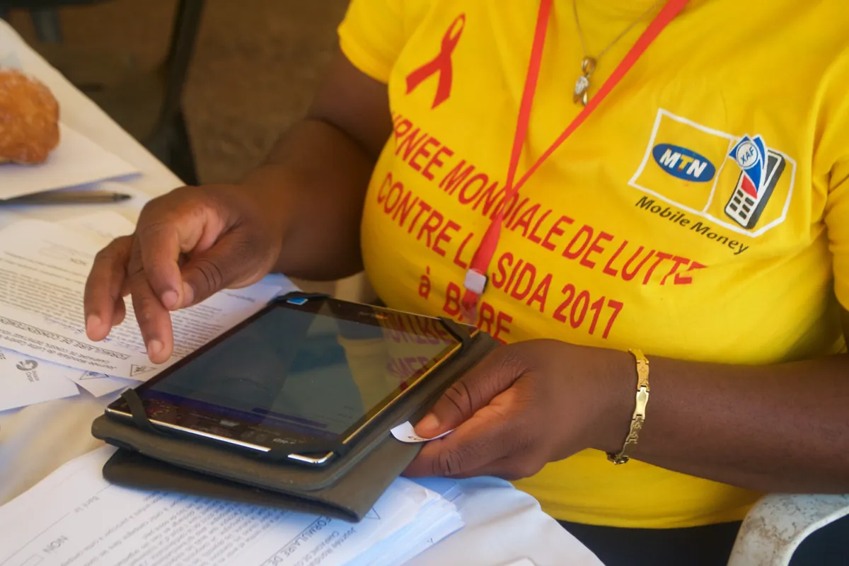 A campaign recorder in a yellow World AIDS Day 2017 T-shirt entering patient information on an Android tablet, with paper forms stacked on the table beside her