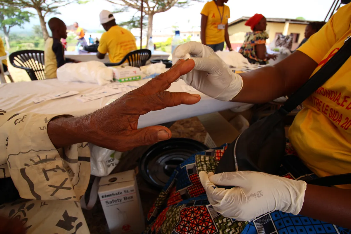 A gloved lab technician performing a finger-prick HIV test for a community member at an outdoor testing table