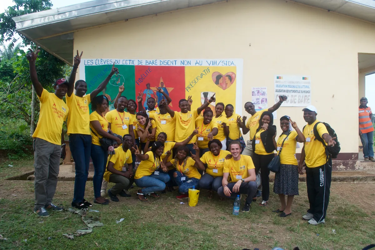 The campaign team in yellow T-shirts posing in front of a student-painted mural reading Les élèves du CETIC de Baré disent non au VIH/SIDA