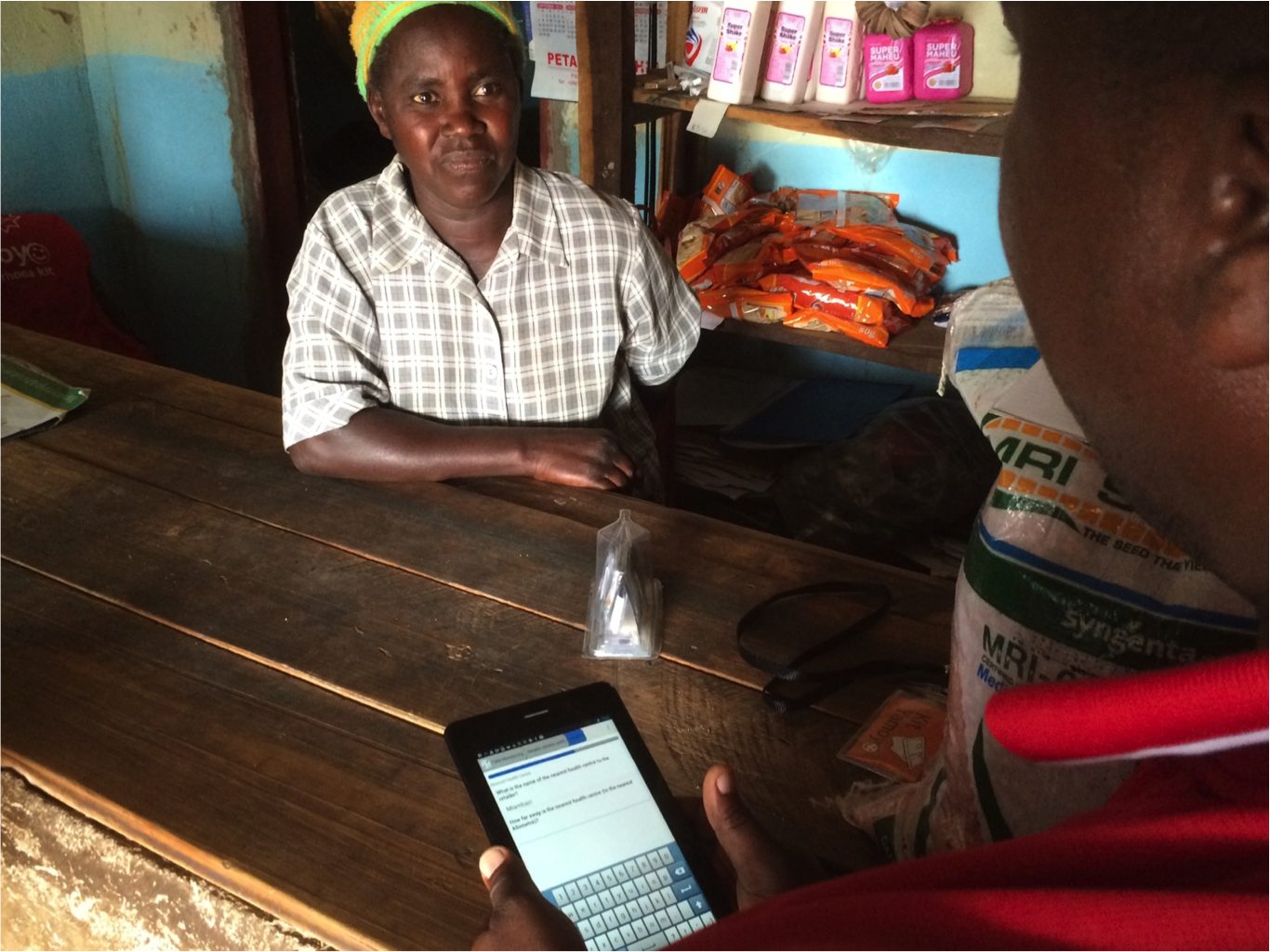 A shopkeeper seated behind a wooden counter in a small store while a customer enters data on a tablet