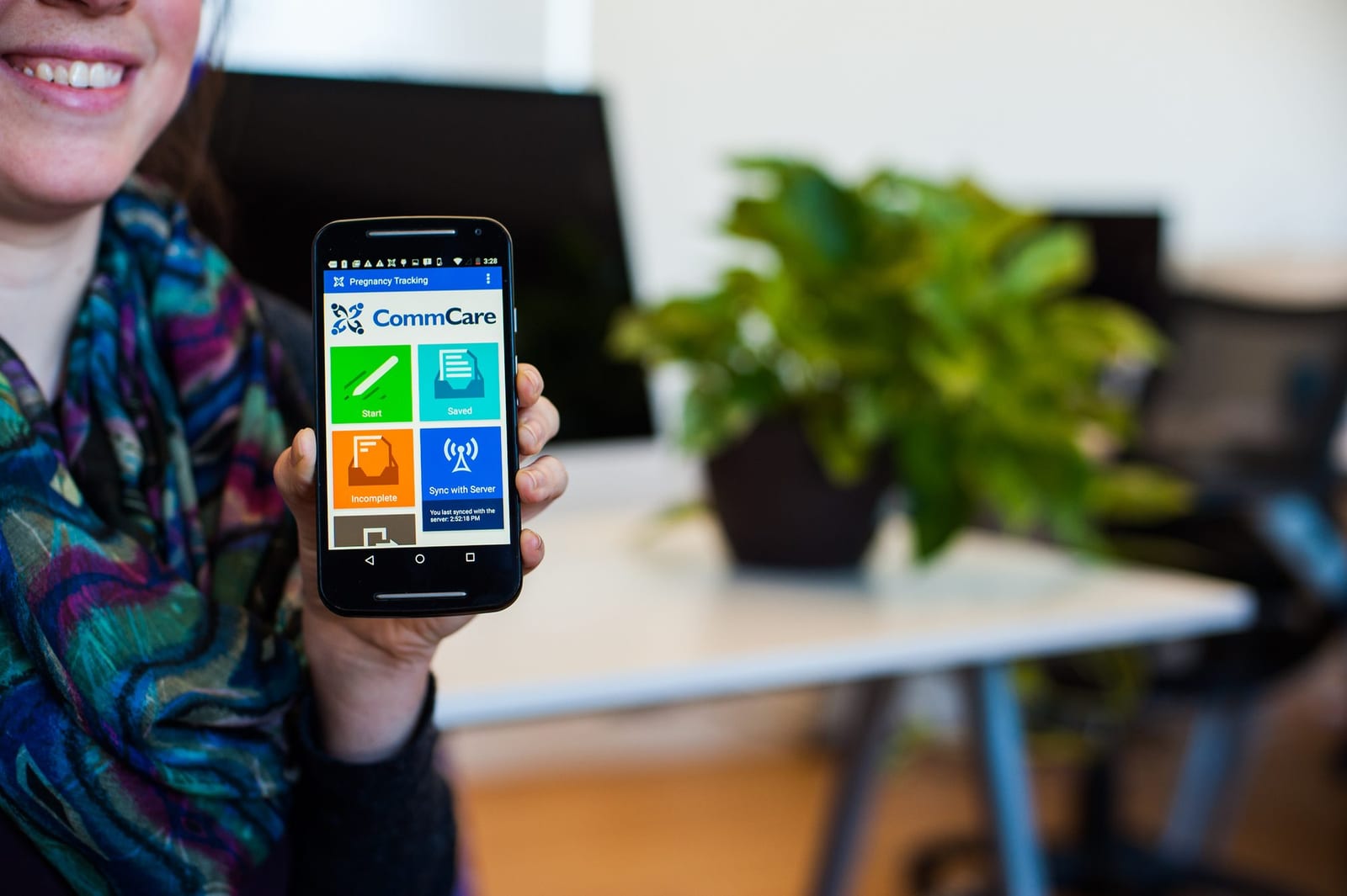 A hand holding a phone displaying the CommCare app home screen in an office